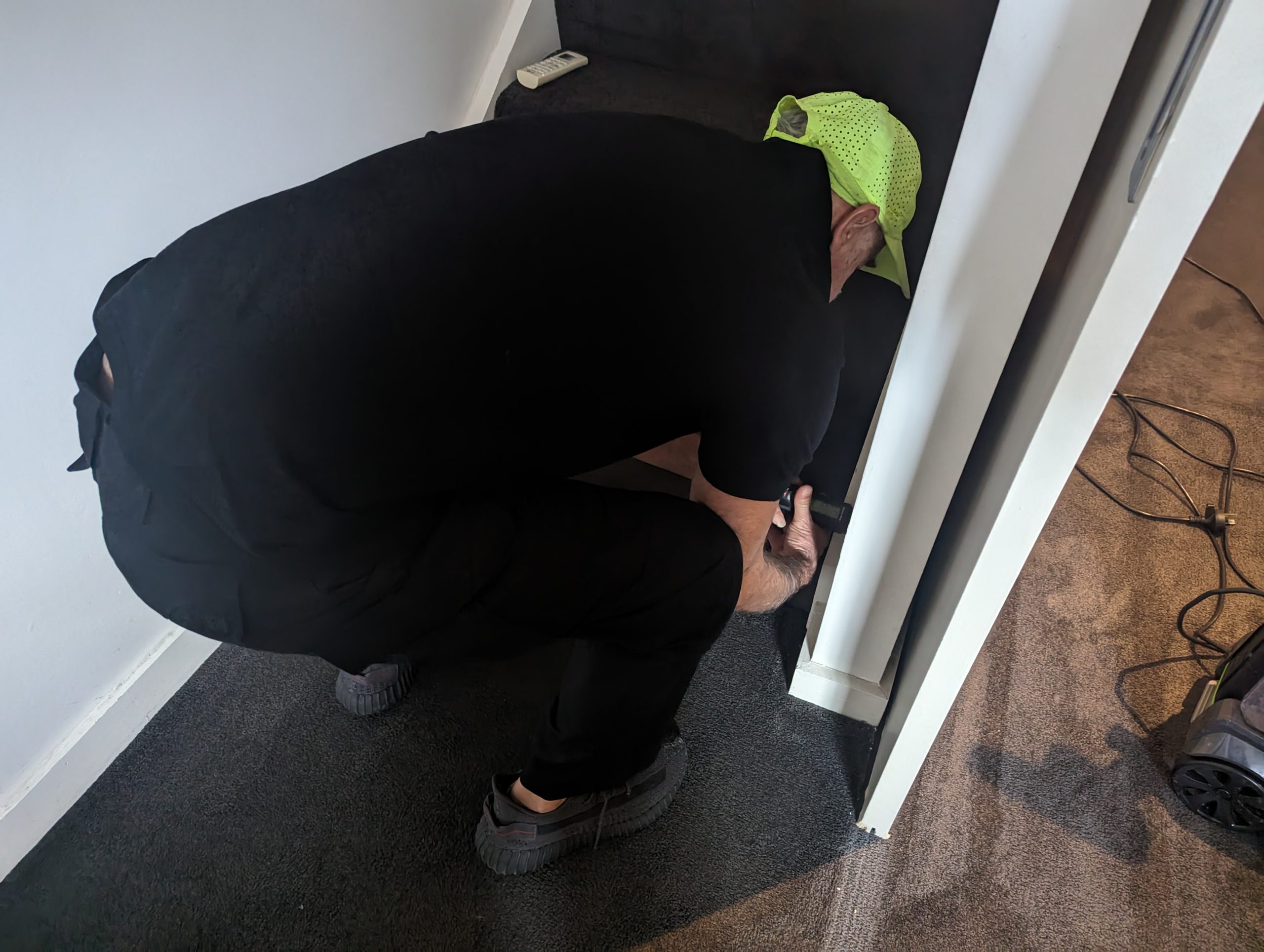 Technician assessing and repairing water damage in a residential setting in Sydney, wearing a bright cap, focused on the base of a wall, with a vacuum cleaner visible nearby.